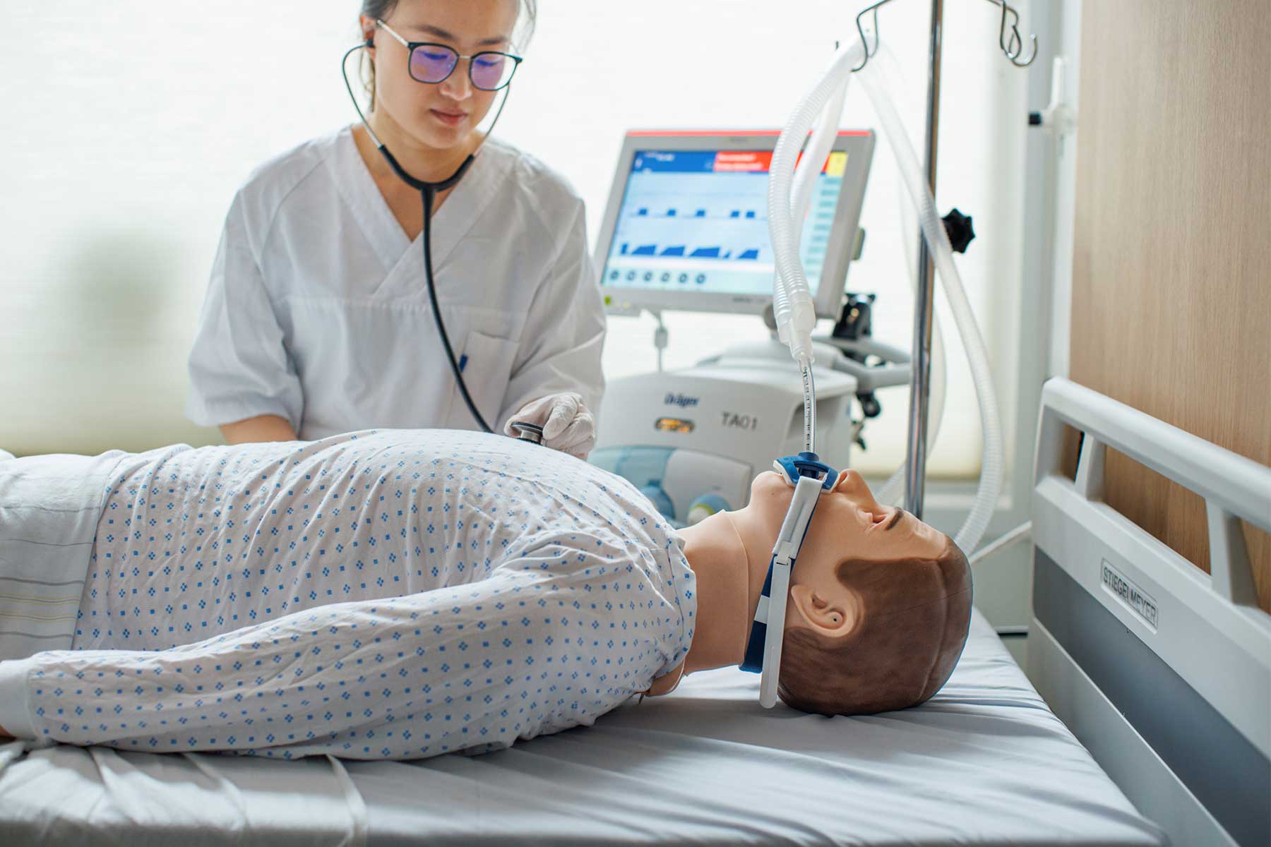 Healthcare worker using a stethoscope to examine a patient lying on a hospital bed with a ventilator.