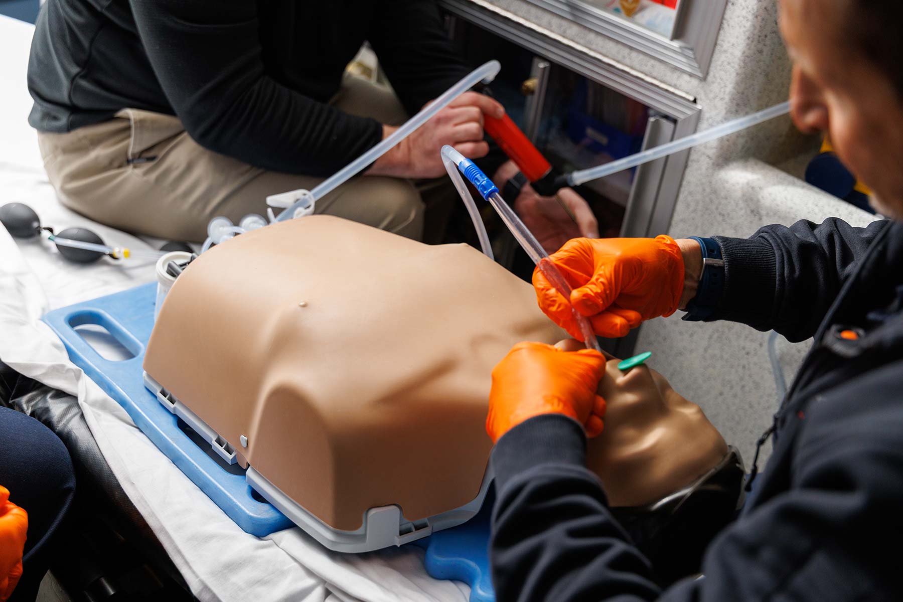 Emergency medical training with two people practicing airway management on a mannequin torso using tubing and a face shield model