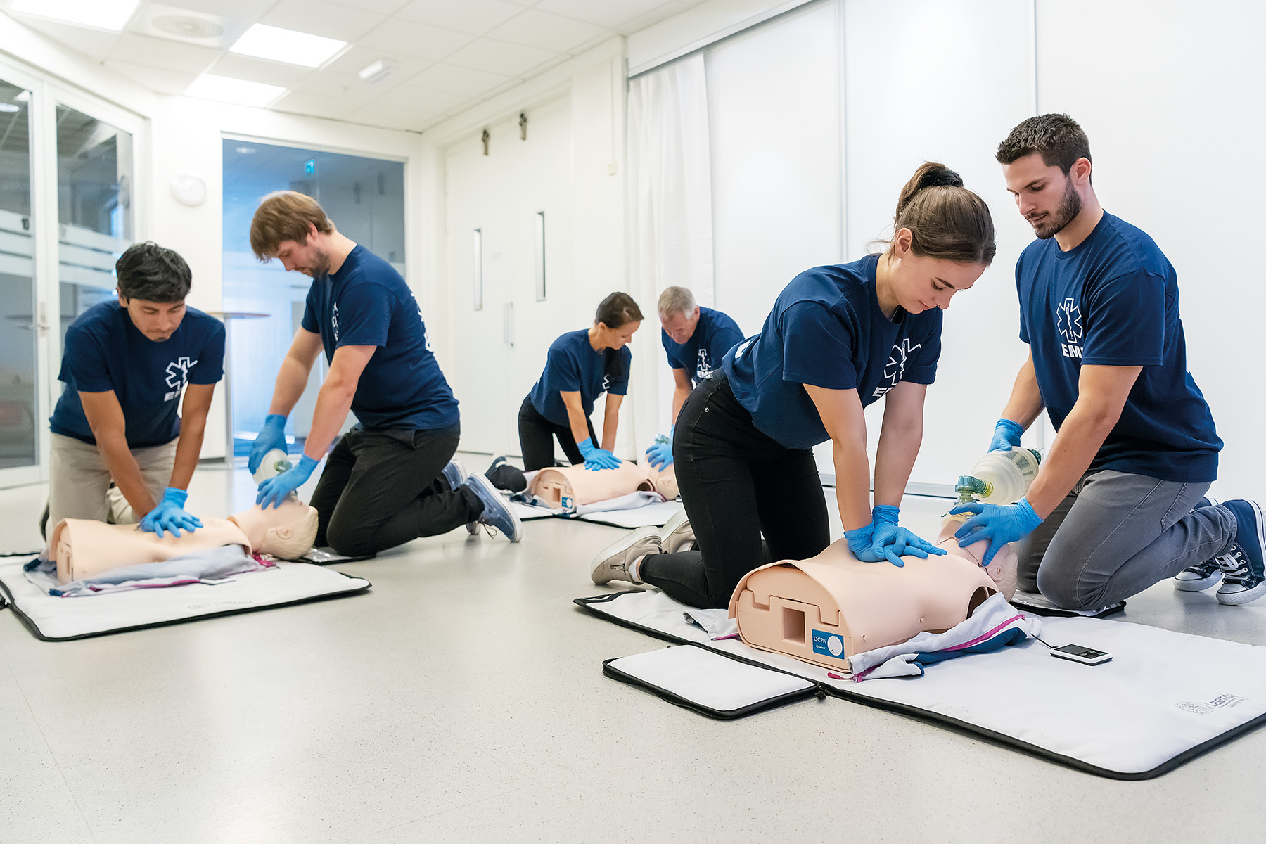 A group of six healthcare trainees or students practicing CPR skills on individual manikins in a bright classroom environment.