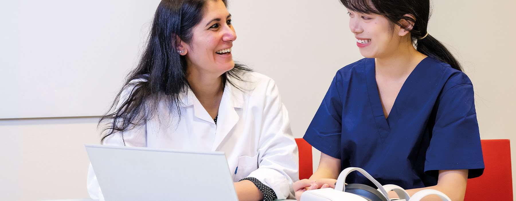 Two women in uniforms, one wearing a white coat and one in a blue nurse uniform, working together on a laptop. Next to the laptop are VR glasses