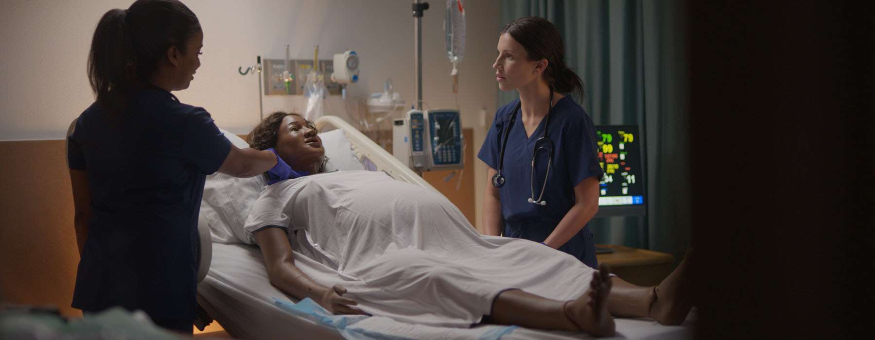 A clinical team performing an initial assessment on a pregnant patient simulator within a realistic, fully equipped hospital simulation room.