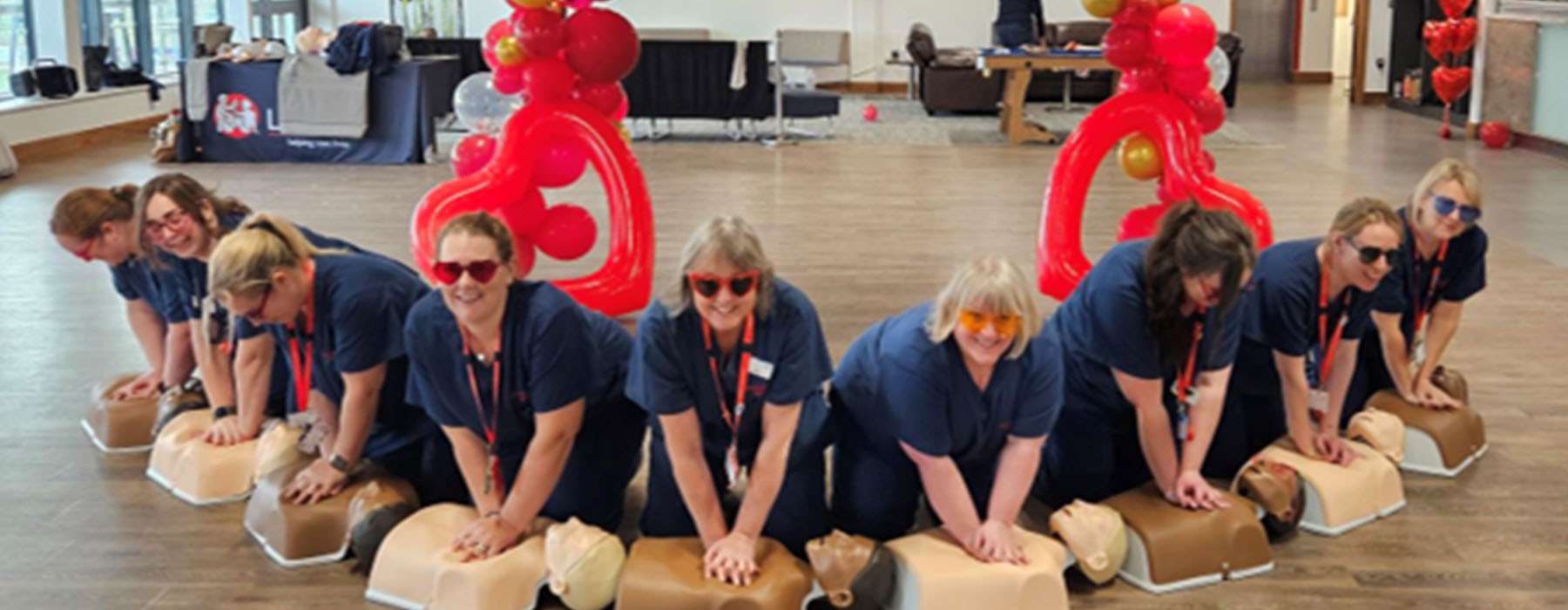 A team of medical staff in a half-circle in front of a red heart balloon structure, doing CPR training with manikins.