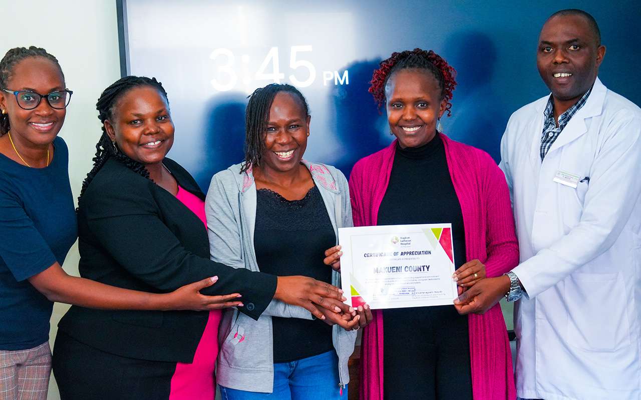 Five people are standing and smiling as a woman in a pink cardigan and a man in a white doctor’s coat hold up a Certificate of Appreciation for Makueni County from Haydom Lutheran Hospital.