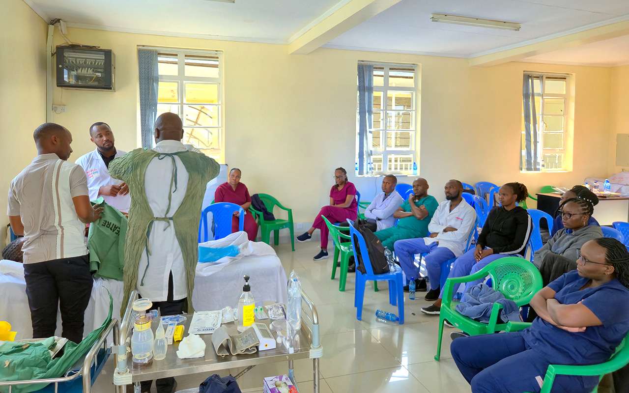 rom a high perspective, a group of about 15 doctors and nurses in blue scrubs are sitting in chairs in a semi-circle and observing a central practical training with simulators
