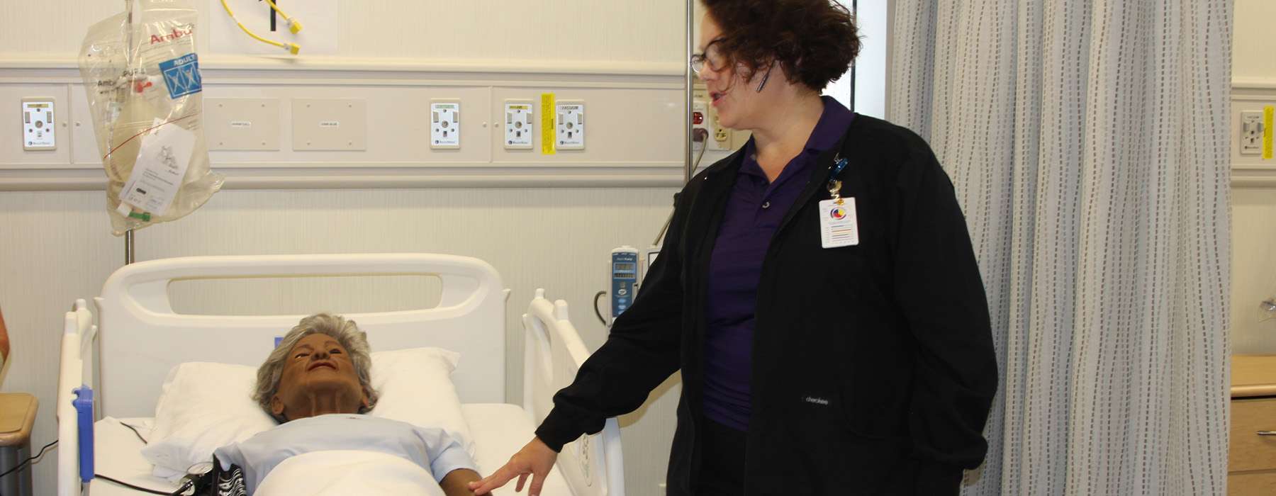 An instructor standing next to a hospital bed with a manikin in it.