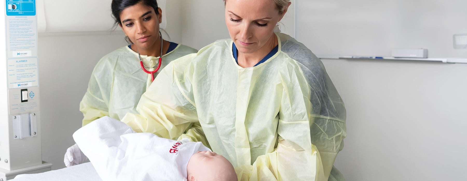 Two nurses wearing yellow operating clothes. One of them is holding a newborn manikin.