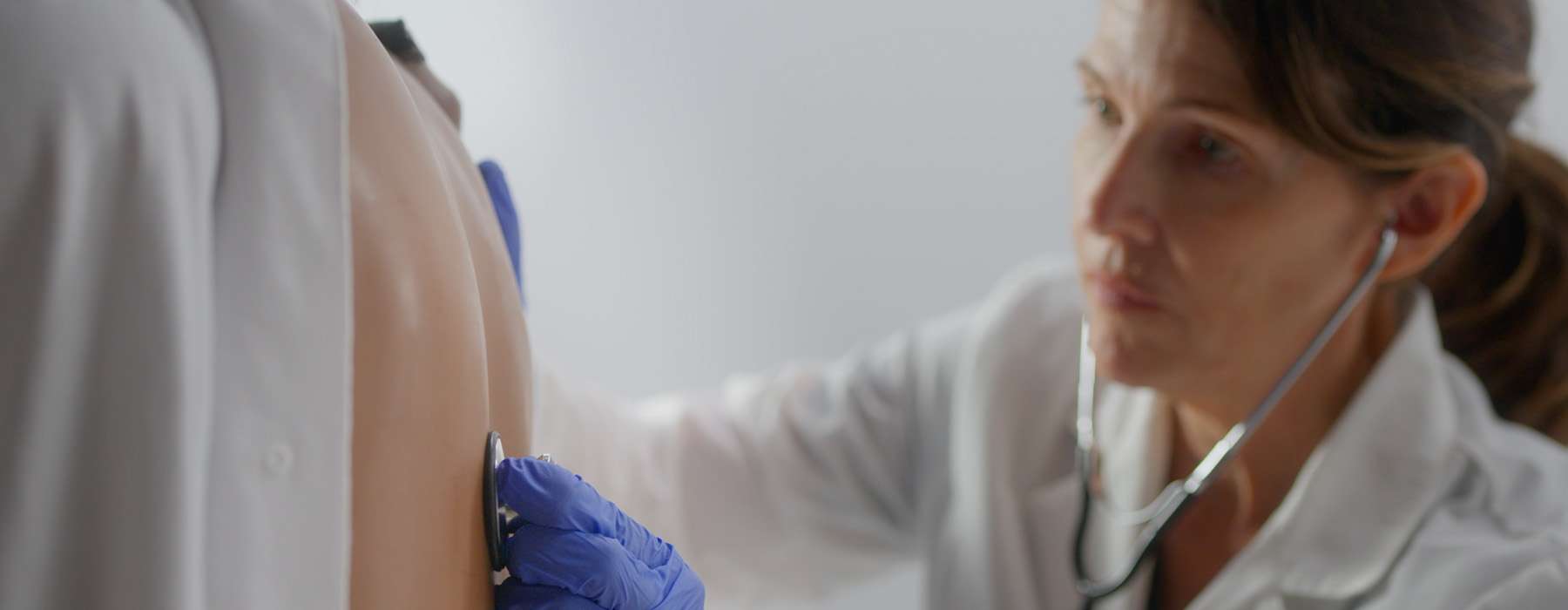 A focused shot of a healthcare professional in a white coat using a stethoscope to assess the back of a patient manikin.