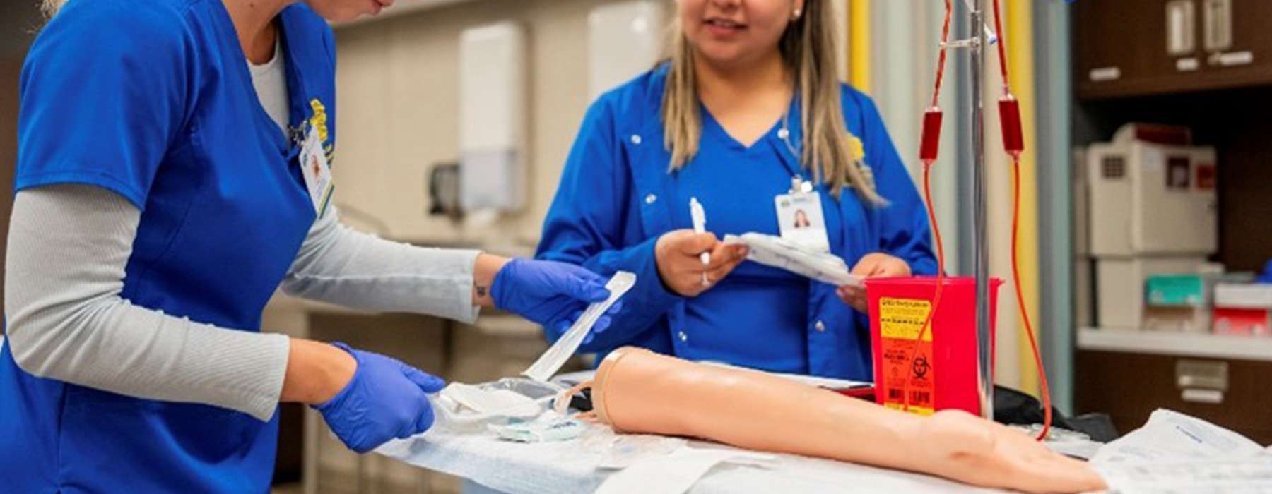 Two nursing students practicing IV catheter preparation and insertion on a task trainer arm.