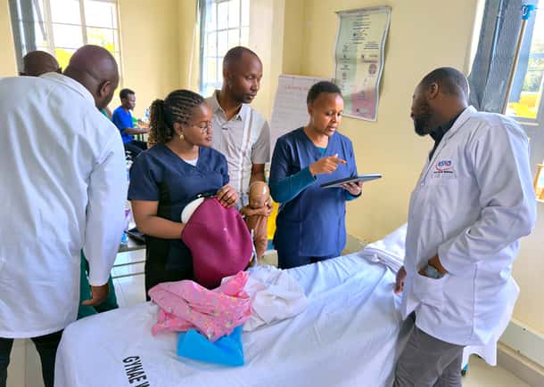 A close-up of a group of five people, including doctors and nurses, around a medical training table and looking down as a woman in blue scrubs holds and explains to them about a MamaNatalie simulator and a newborn simulator. In the background on the wall,